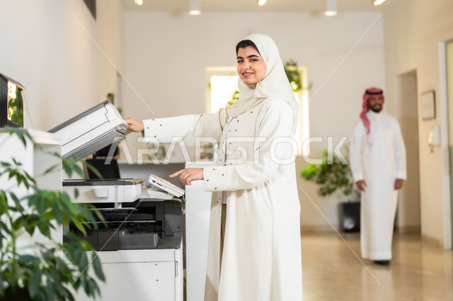 بيئة العمل المريحة في الشركات، مهن و وظائف مكتبية نسائية، موظفة عربية خليجية سعودية محجبة ترتدي عباءة عصرية بيج تقوم بطباعة أوراق العمل الهامة عبر الطابعة الورقية في المكتب، استخدام الأجهزة والآلات التقنية الحديثة في المكاتب