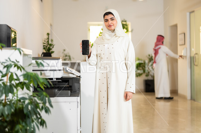 عرض شاشة سوداء فارغة عبر الهاتف المحمول، استخدام الأجهزة التقنية الحديثة في المكاتب، مهن و وظائف مكتبية نسائية، موظفة عربية خليجية سعودية محجبة ترتدي عباءة عصرية بيج تقوم بطباعة أوراق العمل الهامة عبر الطابعة الورقية في المكتب، بيئة العمل المريحة في الشركات