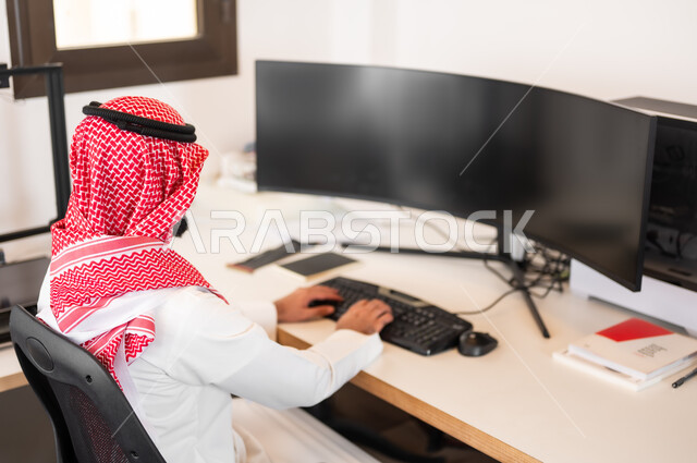 مفهوم إدمان الإنترنت والأجهزة الإلكترونية الحديثة، صورة من الخلف لشاب عربي خليجي سعودي يرتدي الزي التقليدي والشماغ يلعب ألعاب الفيديو على جهاز الكمبيوتر، إجراء بث مباشر (بودكاست) عبر مواقع التواصل الإجتماعي