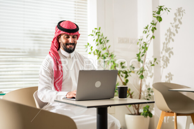 إدارة شؤون الأعمال وتنظيمها عبر الاجهزة التقنية، دمج العمل مع التقنية والتكنولوجيا، مهن ووظائف ادارية مكتبية, رجل اعمال عربي خليجي سعودي يرتدي الزي التقليدي والشماغ يقوم بالعمل على الحاسوب المحمول داخل المكتب