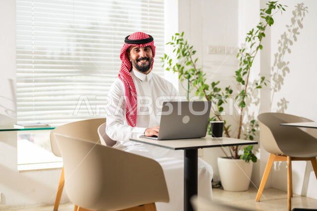 مفهوم إدارة شؤون الأعمال وتنظيمها عبر الاجهزة التقنية، دمج العمل مع التقنية والتكنولوجيا، مهن ووظائف ادارية مكتبية, رجل اعمال عربي خليجي سعودي يرتدي الزي التقليدي والشماغ يقوم بالعمل على الحاسوب المحمول داخل المكتب