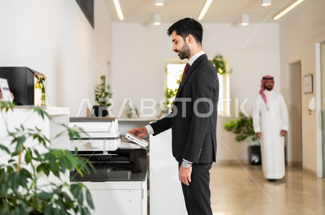 العمل في الشركات الخليجية الراقية, استخدام الأجهزة والآلات التقنية الحديثة في المكاتب، مهن و وظائف مكتبية، صورة من الجانب لموظف عربي خليجي سعودي يرتدي الزي الرسمي يقوم بطباعة أوراق العمل الهامة عبر الطابعة الورقية في المكتب