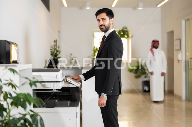 الوقوف بثقة والنظر إلى الكاميرا, العمل في الشركات الخليجية الراقية, استخدام الأجهزة والآلات التقنية الحديثة في المكاتب، مهن و وظائف مكتبية، صورة من الجانب لموظف عربي خليجي سعودي يرتدي الزي الرسمي يقوم بطباعة أوراق العمل الهامة عبر الطابعة الورقية في المكتب