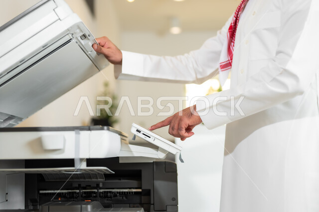 العمل في الشركات الخليجية الراقية, استخدام الأجهزة والآلات التقنية الحديثة في المكاتب، مهن و وظائف مكتبية، صورة من الجانب لموظف عربي خليجي سعودي يرتدي الثوب التقليدي والشماغ يقوم بطباعة أوراق العمل الهامة عبر الطابعة الورقية في المكتب