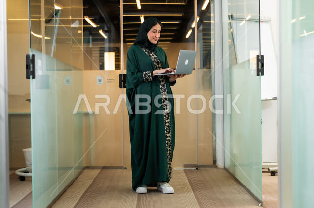 مهن و وظائف نسائية، استخدام التقنية والتكنولوجيا لإنجاز المهام والمشاريع، ، سيدة أعمال عربية خليجية سعودية مبتسمة محجبة ترتدي عباءة زيتي تستخدم جهاز الحاسوب لانجاز العمل في مقر الشركة