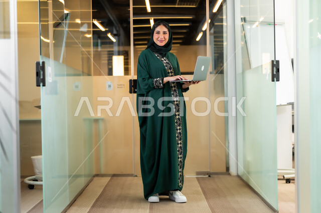 Using technology and technology to accomplish tasks and projects, women’s jobs and professions, a smiling Saudi Arabian Gulf businesswoman, veiled and wearing an olive abaya, uses a computer to complete work at the company’s headquarters