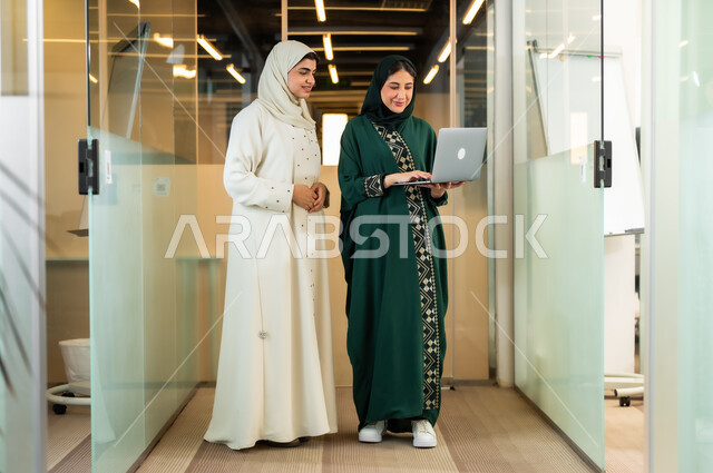 تطوير ومتابعة العمل في الشركات السعودية، مهن و وظائف نسائية, موظفتان عربيتان خليجتان ترتديان عباءات تقليدية عصرية بالوان مختلفة استخدام جهاز الحاسوب في مقر الشركة,استخدام الوسائل التقنية الحديثة والمتطورة لإنجاز المهام