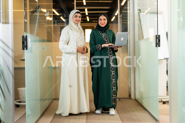 متابعة وتطوير العمل في الشركات السعودية، مهن و وظائف نسائية, موظفتان عربيتان خليجتان ترتديان عباءات تقليدية عصرية بالوان مختلفة استخدام جهاز الحاسوب في مقر الشركة,استخدام الوسائل التقنية الحديثة والمتطورة لإنجاز المهام