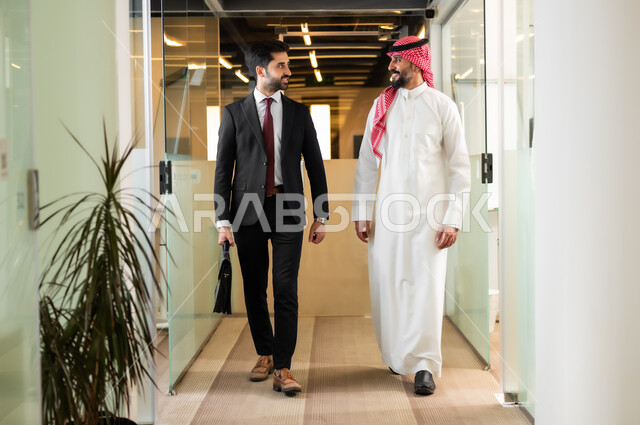 مناقشة الخطط وشرح مهام الاعمال، حوارات ونقاشتات عمل ، رجل عربي سعودي خليجي يرتدي الثوب التقليدي والشماغ مع رجل بالبدلة الرسمية  يمشيان داخل مقر الشركة، بيئة العمل المريحة في الشركات السعودية