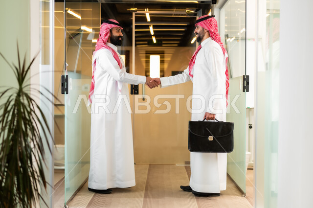 التحية والسلام والاتفاق على صفقات عمل ناجحة, العمل في الشركات الخليجية الراقية, رجلا أعمال عربيان خليجيان سعوديان يرتديان الثوب التقليدي والشماغ يقومان بالمصافحة بتعابير السرور يقفون داخل مقر الشركة
