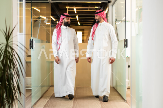 بيئة العمل المريحة في الشركات السعودية، مناقشة الخطط وشرح مهام الاعمال، زملاء عمل عربيان سعوديان خليجيان يرتديان الثوب التقليدي والشماغ يقومان بالمشي داخل مقر الشركة، الاتفاق على المشاريع التجارية