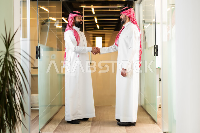 العمل في الشركات الخليجية الراقية, التحية والسلام والاتفاق على صفقات عمل ناجحة, رجلا أعمال عربيان خليجيان سعوديان يرتديان الثوب التقليدي والشماغ يقومان بالمصافحة بتعابير السرور داخل مقر الشركة