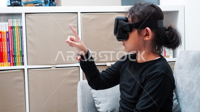 رؤية رسومات متحركة ولعب ألعاب الفيديو، الإندماج والتفاعل مع العالم الخيالي، قضاء أوقات ممتعة بمتابعة أفلام الكرتون عبر نظارة الواقع الافتراضي، صورة مقربة لفتاة عربية خليجية سعودية ترتدي نظارات ثلاثية الأبعاد تقوم بالإشارة الى شيئ ما