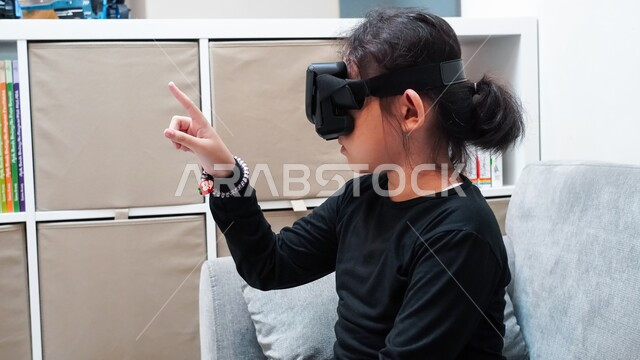 مشاهدة رسومات متحركة ولعب ألعاب الفيديو، الإندماج والتفاعل مع العالم الخيالي، قضاء أوقات ممتعة بمتابعة أفلام الكرتون عبر نظارة الواقع الافتراضي، صورة مقربة لفتاة عربية خليجية سعودية ترتدي نظارات ثلاثية الأبعاد تقوم بالإشارة الى شيئ ما