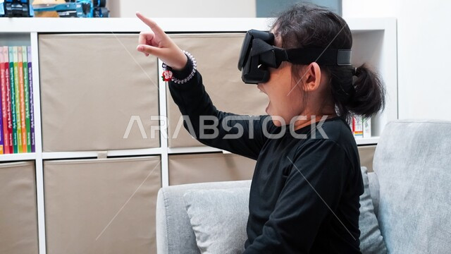 لعب ألعاب الفيديو ومشاهدة رسومات متحركة، الإندماج والتفاعل مع العالم الخيالي، قضاء أوقات ممتعة بمتابعة أفلام الكرتون عبر نظارة الواقع الافتراضي، صورة مقربة لفتاة عربية خليجية سعودية ترتدي نظارات ثلاثية الأبعاد تقوم بالإشارة الى شيئ ما