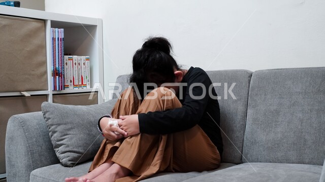 الدخول بحالة إكتئاب بسبب شيء ما، ايماءات الحزن والشعور بالوحدة والقلق، فتاة عربية خليجية سعودية تجلس على أريكة رمادية في غرفة المعيشة منطوية على نفسها بتعابير الإحباط