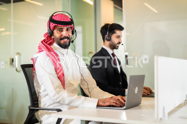الاجابة على الاستعلامات والاستفسارات, النظر إلى الكاميرا بإيماءات السعادة، بيئة العمل المريحة في الشركات الراقية, رجل عربي سعودي خليجي مبتسمان يرتديان سماعة الرأس يقومان في التواصل مع الزبائن لحل المشكلات
