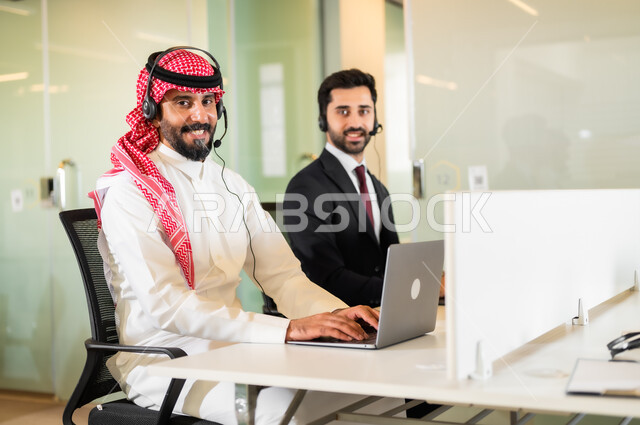 النظر إلى الكاميرا بإيماءات السعادة، بيئة العمل المريحة في الشركات الراقية, رجل عربي سعودي خليجي مبتسمان يرتديان سماعة الرأس يقومان في التواصل مع الزبائن لحل المشكلات, الاجابة على الاستعلامات والاستفسارات
