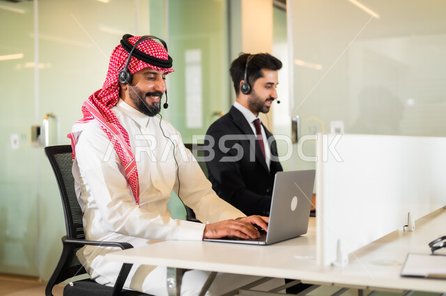 العمل على الجهزة الحاسوب في مقر الشركة, الإجابات على الأسئلة والاستفسارات, رجل عربي سعودي خليجي وشاب يرتدي البدلة الرسمية مبتسمان يرتديان سماعة الرأس يقومان في التواصل مع الزبائن لحل المشكلات والشكاوي, مفهوم المشاركة و التعاون في العمل