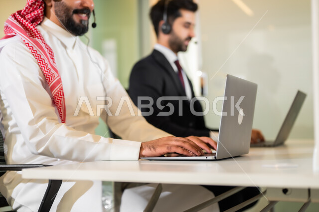 The concept of participation and cooperation at work, answers to questions and inquiries, a Saudi Gulf Arab man with a young man wearing a formal suit, two smiling men wearing headphones, communicating with customers to solve problems and complaints, working on computers at the company’s headquarters.
