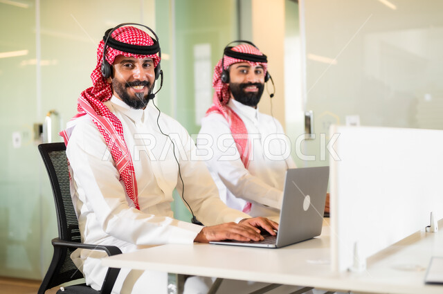 الاجابة على الاستعلامات والاستفسارات، بيئة العمل المريحة في الشركات الراقية, موظفان عربيان سعوديان خليجيان مبتسمان يرتديان الزي التقليدي والشماغ و سماعة الرأس يقومان في التواصل مع الزبائن لحل المشكلات, النظر إلى الكاميرا بإيماءات السعادة