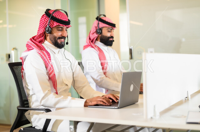 الإجابة على استفسارات العملاء, بيئة العمل المريحة في الشركات الراقية, موظفان عربيان سعوديان خليجيان مبتسمان يرتديان الزي التقليدي والشماغ وسماعة الرأس يقومان في التواصل مع الزبائن لحل المشكلات والشكاوي, العمل على الجهز الحاسوب في مقر الشركة