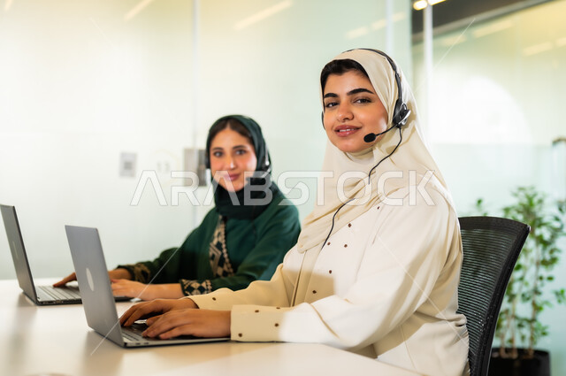 العمل على جهاز الحاسوب في مقر الشركة, وظائف ومهن مكتبية نسائية, الإجابة عن الاستعلامات والاستفسارات, سيدتان عربيتان سعوديتان خليجيتان محجبتان مبتسمتان ترتديان العباءة وسماعة الرأس يقومان في التواصل مع الزبائن لحل المشكلات, النظرإلى الكاميرا بإيماءات السرور