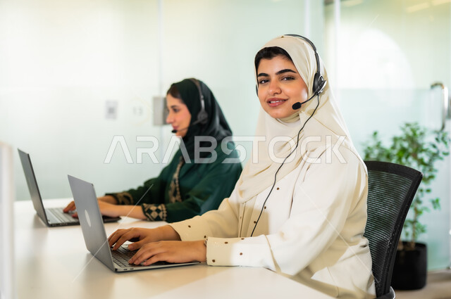 وظائف ومهن مكتبية نسائية, الإجابة عن الاستعلامات والاستفسارات,سيدتان عربيتان سعوديتان خليجيتان محجبتان مبتسمتان ترتديان العباءة وسماعة الرأس يقومان في التواصل مع الزبائن لحل المشكلات, العمل على جهاز الحاسوب في مقر الشركة, النظرإلى الكاميرا بإيماءات السرور