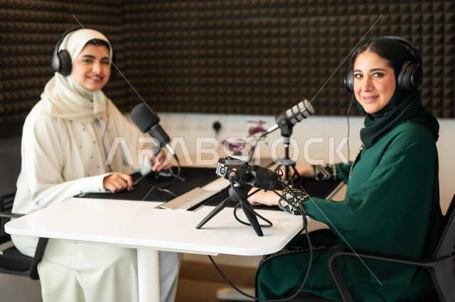 Expressions of enthusiasm and pleasure, use of modern technical devices, two veiled Saudi Arabian Gulf women, smiling, wearing abayas and headphones, working in the field of audio broadcasting, dialogues and discussions, inquiries and enquiries, looking at the camera with happy gestures.