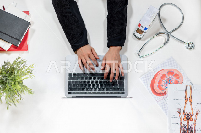  تصوير مقرب من الأعلى , لطبيبة عربية سعودية لديها خبره في طب الباطنية , طبيبة باطنية تعمل من خلال اللابتوب على مكتب أبيض , وظيفه ومهن , بيئة عمل , خلفية بيضاء .