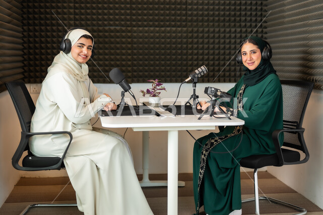 النظر إلى الكاميرا بايماءات السرور، سيدتان عربيتان خليجيتان محجبتان مبتسمتان ترديان العباءة وسماعة الرأس, مقابلة عمل داخل الاستديو, معدات وأجهزة تسجيلات الصوت، اجراء بث صوتي مباشر (بودكاست) عبر برامج التواصل الاجتماعي