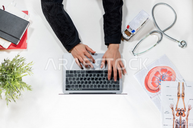  تصوير مقرب من الأعلى , لطبيبة عربية سعودية لديها خبره في طب الباطنية , طبيبة باطنية تعمل من خلال اللابتوب على مكتب أبيض , وظيفه ومهن , بيئة عمل , خلفية بيضاء .