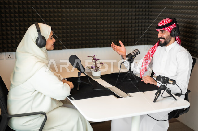 ايماءات السعادة والفرح، رجل عربي سعودي خليجي يرتدي الثوب التقليدي والشماغ  يرفع يده للأعلى مع شابة بالعباءة والحجاب في مقابلة عمل داخل مقر الاستديو، معدات وأجهزة تسجيلات الصوت، اجراء بث صوتي مباشر (بودكاست) عبر برامج التواصل الاجتماعي، ايماءات الحماس والتفاعل
