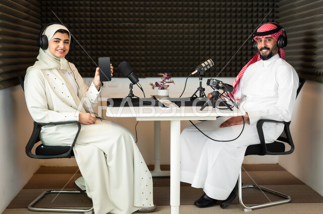 Looking at the camera with happy gestures, a Saudi Gulf Arab man wearing a traditional thobe and shemagh with a young woman in an abaya and hijab holding a black blank screen phone in her hand in a job interview inside the studio headquarters, audio recording equipment and devices, conducting a live audio broadcast (podcast) via social media programs