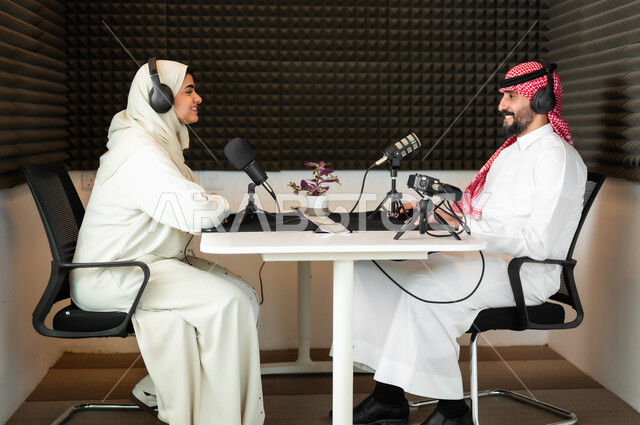 استديو تسجيل صوتي،  رجل عربي سعودي خليجي يرتدي الثوب التقليدي والشماغ مع شابة بالعباءة والحجاب في مقابلة عمل داخل مقر الاستديو، تعابير الفرح والسرور، معدات وأجهزة تسجيلات الصوت، اجراء بث صوتي مباشر (بودكاست) عبر برامج التواصل الاجتماعي، ايماءات الحماس والتفاعل