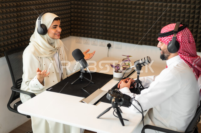 ايماءات تدل على الحماس، رجل عربي سعودي خليجي يرتدي الثوب التقليدي والشماغ مع شابة بالعباءة والحجاب في مقابلة عمل داخل مقر الاستديو، معدات وأجهزة تسجيلات الصوت، اجراء بث صوتي مباشر ( بودكاست ) عبر برامج التواصل الاجتماعي، تعابير السعادة والسرور