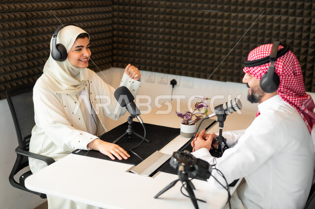 تعابير السعادة والسرو، مقابلة عمل داخل مقر الشركة، رجل عربي سعودي خليجي يرتدي الثوب التقليدي والشماغ مع شابة بالعباءة والحجاب ترفع يدها للأعلى في مقابلة عمل داخل مقر الاستديو،  معدات وأجهزة تسجيلات الصوت، اجراء بث صوتي مباشر (بودكاست) عبر برامج التواصل الاجتماعي