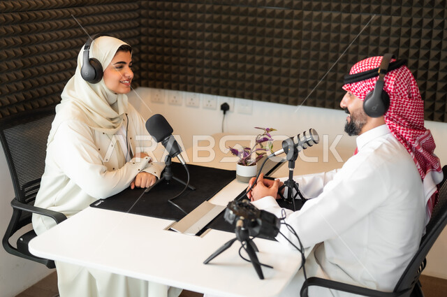 ايماءات الفرح والسرور، رجل عربي سعودي خليجي يرتدي الثوب التقليدي والشماغ مع شابة بالعباءة والحجاب في مقابلة عمل داخل مقر الاستديو، معدات وأجهزة تسجيلات الصوت، اجراء بث صوتي مباشر (بودكاست) عبر برامج التواصل الاجتماعي