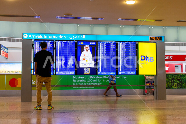 الفن الهندسي المتميز والديكور الداخلي للمطارات، لافتة الكترونية ارشادية، مراقبة موعد رحلات السفر، صورة من الخلف لمسافر يقف بجانب لوحة القادمون داخل مطار دبي الدولي بالإمارات العربية المتحدة