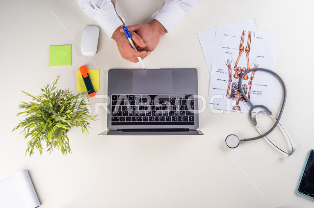  تصوير مقرب من الأعلى , لطبيب عربي سعودي لديه خبره في طب الباطنية , طبيب باطنية يعمل من خلال اللابتوب على مكتب أبيض , وظيفه ومهن , بيئة عمل , خلفية بيضاء .