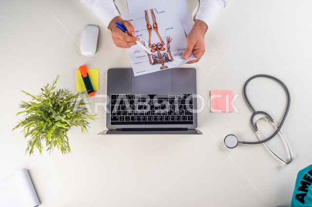  تصوير مقرب من الأعلى , لطبيب عربي سعودي لديه خبره في طب الباطنية , طبيب باطنية يعمل من خلال اللابتوب على مكتب أبيض , وظيفه ومهن , بيئة عمل , خلفية بيضاء .