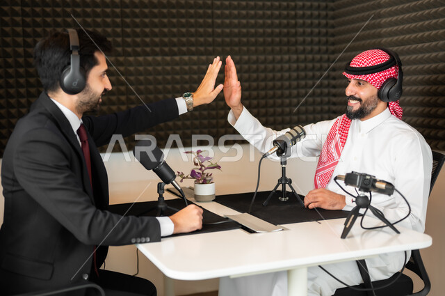 تعابير السعادة والسرور، ايماءات الحماس والتفاعل، رجل عربي سعودي خليجي يرتدي الثوب والشماغ التقليدي مع رجل بالبدلة الرسمية يضربان كيفهما ببعض، معدات وأجهزة تسجيلات الصوت، اجراء بث صوتي مباشر (بودكاست) عبر برامج التواصل الاجتماعي