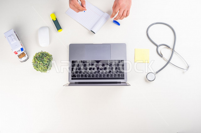  تصوير مقرب من الأعلى , لطبيب عربي سعودي لديه خبره في طب الباطنية , طبيب باطنية يعمل من خلال اللابتوب على مكتب أبيض , وظيفه ومهن , بيئة عمل , خلفية بيضاء .