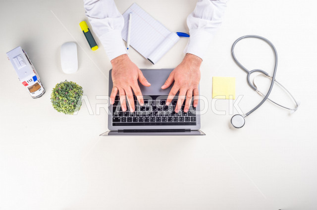  تصوير مقرب من الأعلى , لطبيب عربي سعودي لديه خبره في طب الباطنية , طبيب باطنية يعمل من خلال اللابتوب على مكتب أبيض , وظيفه ومهن , بيئة عمل , خلفية بيضاء .