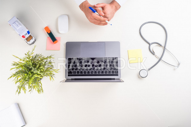  تصوير مقرب من الأعلى , لطبيب عربي سعودي لديه خبره في طب الباطنية , طبيب باطنية يعمل من خلال اللابتوب على مكتب أبيض , وظيفه ومهن , بيئة عمل , خلفية بيضاء .