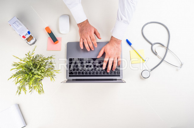  تصوير مقرب من الأعلى , لطبيب عربي سعودي لديه خبره في طب الباطنية , طبيب باطنية يعمل من خلال اللابتوب على مكتب أبيض , وظيفه ومهن , بيئة عمل , خلفية بيضاء .