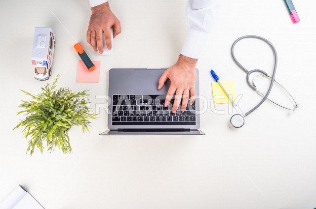  تصوير مقرب من الأعلى , لطبيب عربي سعودي لديه خبره في طب الباطنية , طبيب باطنية يعمل من خلال اللابتوب على مكتب أبيض , وظيفه ومهن , بيئة عمل , خلفية بيضاء .