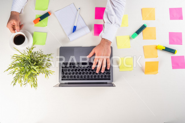 صورة مقربة من الأعلى , لموظف سعودي عربي يعمل من خلال اللابتوب على مكتب أبيض , مهن ووظيفة , بيئة عمل , خلفية بيضاء .