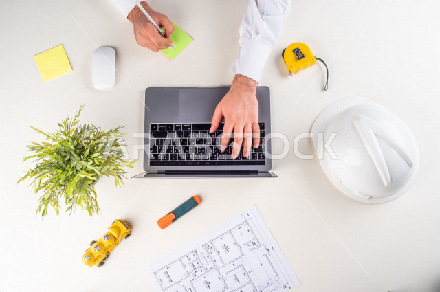  تصوير مقرب من الأعلى , لمهندس معماري خليجي , مهندس يعمل على تصميم العمارات من خلال اللابتوب على مكتب أبيض , وظيفه ومهن , بيئة عمل , خلفية بيضاء .