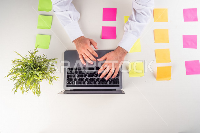 صورة مقربة من الأعلى , لموظف سعودي عربي يعمل من خلال اللابتوب على مكتب أبيض , مهن ووظيفة , بيئة عمل , خلفية بيضاء .